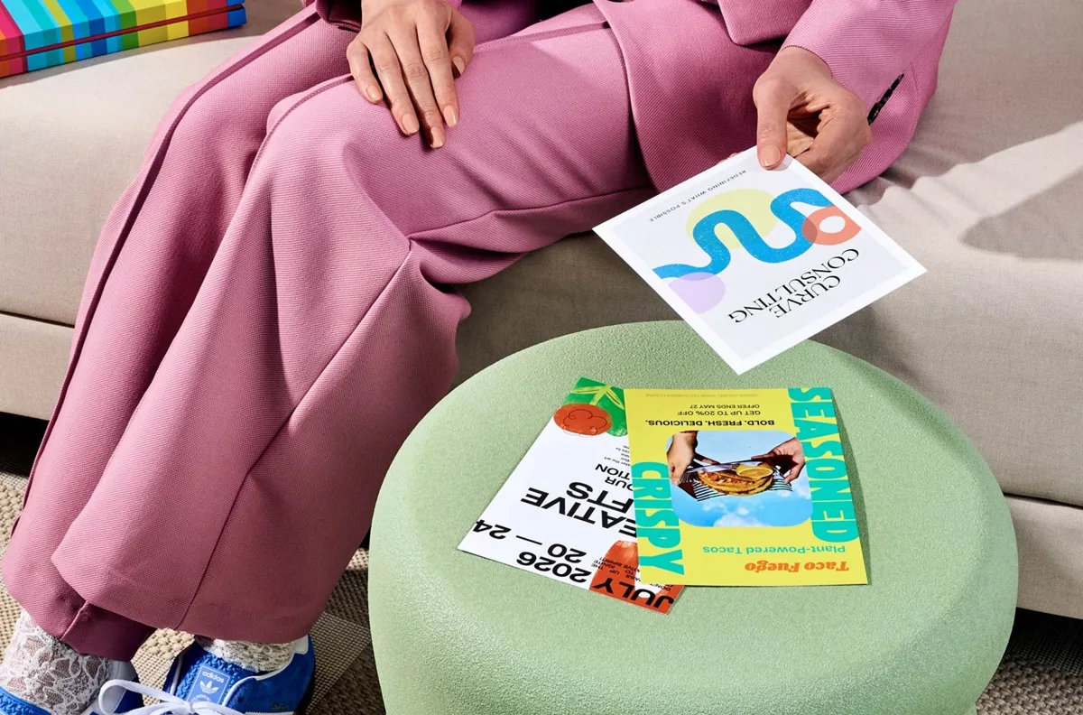 A person on a sofa holding a square postcard, with two more postcards on a stool